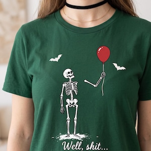 May include: A dark green t-shirt featuring a graphic of a skeleton looking up at a detached skeletal arm holding a red balloon. Two white bats are depicted in the background. The text "Well, shit..." is printed in white at the bottom of the graphic.