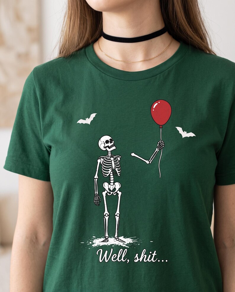 May include: A dark green t-shirt featuring a graphic of a skeleton looking up at a detached skeletal arm holding a red balloon. Two white bats are depicted in the background. The text "Well, shit..." is printed in white at the bottom of the graphic.