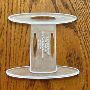 May include: A clear acrylic spool-shaped tool with two oval cutouts on the central stem. The text printed on the stem reads: The Binding Bobbin, TQM Products, 2025-The Quilters Mercantile, Made in the USA, tqmrulers.com. The object has a flat base.