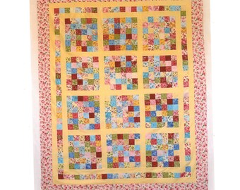 May include: A rectangular patchwork quilt featuring twelve square blocks of multi-colored floral and solid fabric squares separated by yellow sashing. The quilt is framed by a wide border of pink and white floral fabric on a light background.