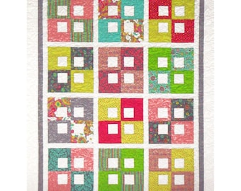 May include: A quilt featuring a grid of twelve blocks, each composed of four smaller squares with a central white cutout. The blocks display a variety of colorful floral, striped, and dotted patterns in shades of pink, green, yellow, and teal, all framed by a solid gray border and a white quilted edge.