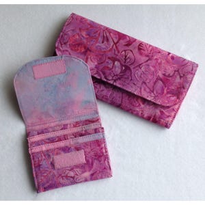 May include: A rectangular wallet and a smaller folding wallet, both featuring a vibrant pink and purple floral batik pattern. The smaller wallet is open, revealing multiple card slots and a rectangular hook-and-loop fastener on the flap.
