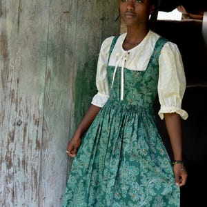 May include: A floor-length green dress featuring a detailed floral damask pattern, paired with a cream-colored blouse that has short puff sleeves and a front tie closure. The dress has a fitted bodice and a gathered, full skirt with a defined waistline.
