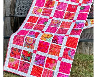 May include: A patchwork quilt featuring a grid of square fabric patches in various shades of pink, red, and orange, with floral, polka dot, and geometric patterns. The squares are separated by white fabric sashing and finished with white stitching.