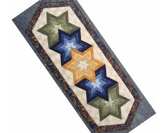 May include: A rectangular quilted table runner featuring a central row of five overlapping star shapes in shades of olive green, navy blue, and golden yellow. The stars are set against a cream-colored background with a dark blue floral border.