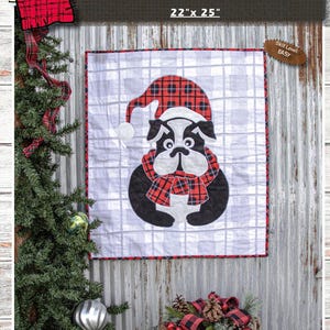 May include: A square quilt pattern titled Cutest Brutus measuring 22 inches by 25 inches. The design features a black and white bulldog wearing a red and black buffalo plaid hat and scarf against a light gray and white checkered background.
