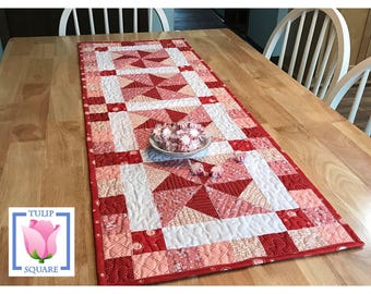 May include: A rectangular quilted table runner featuring a patchwork design in shades of red, pink, and white. The pattern includes pinwheel blocks and solid squares with intricate stitching. A small bowl of peppermint candies sits in the center.