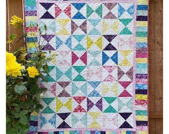 May include: A rectangular quilt featuring a repeating bowtie pattern in various colors including purple, pink, yellow, blue, and green, set against a white background. The quilt has a multi-colored striped border and a thin pink inner border.
