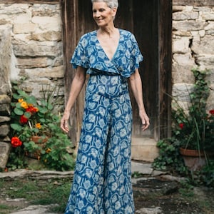 May include: A floor-length jumpsuit in a deep blue hue featuring a repeating white floral pattern. The garment has a V-neckline, short flutter sleeves, and wide-leg trousers that drape to the floor, creating a fluid and continuous silhouette.