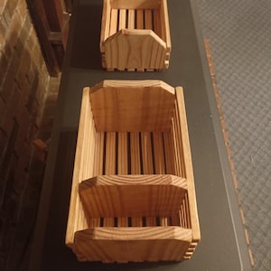 May include: Two rectangular wooden crates constructed from light brown slats. The smaller crate is open, while the larger crate features a central wooden divider, creating two distinct compartments within the slatted structure of the container.