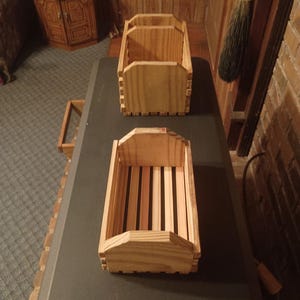 May include: Two rectangular wooden crates constructed from light-colored slats with vertical end panels featuring angled top corners. The crates have an open-top design with visible grain patterns and are arranged one behind the other on a surface.