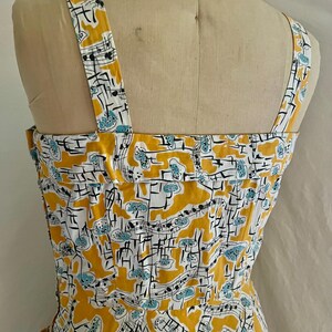 K&ouml;nnte beinhalten: Ein Vintage-Kleid mit einem Notenmuster. Das Kleid hat ein gelbes, wei&szlig;es und blaues Farbschema. Die Tr&auml;ger und das Oberteil sind sichtbar und zeigen das Design. Das Kleid wird auf einer Schaufensterpuppe pr&auml;sentiert.