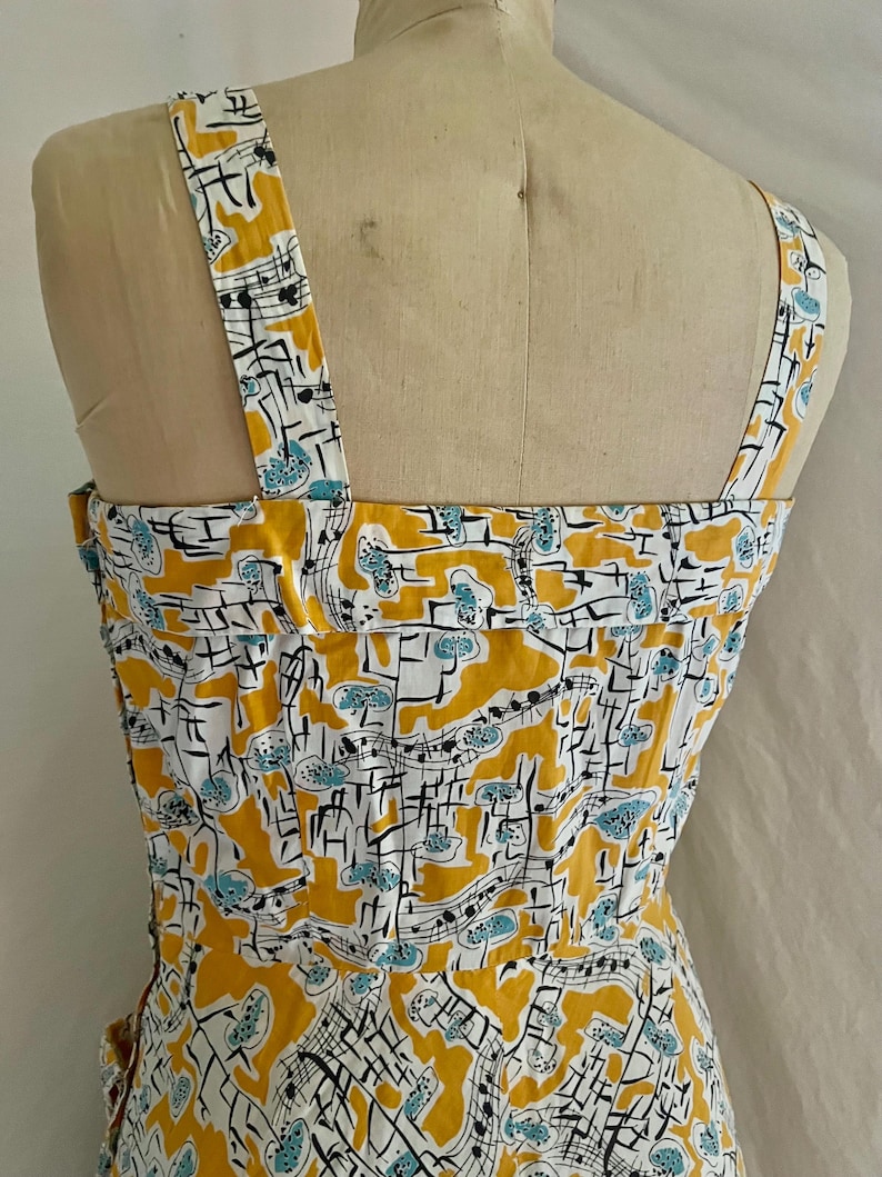 K&ouml;nnte beinhalten: Ein Vintage-Kleid mit einem Notenmuster. Das Kleid hat ein gelbes, wei&szlig;es und blaues Farbschema. Die Tr&auml;ger und das Oberteil sind sichtbar und zeigen das Design. Das Kleid wird auf einer Schaufensterpuppe pr&auml;sentiert.