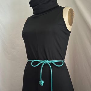 Può includere: Un abito nero senza maniche con collo alto a lupetto. Una cintura turchese è annodata in vita, con nodi decorativi alle estremità. L'abito è esposto su un manichino.