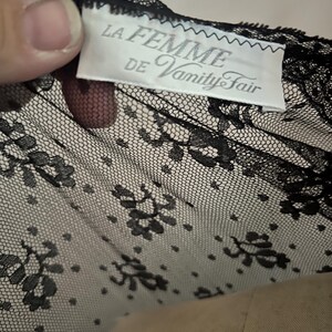 May include: Black lace fabric with a floral pattern. The tag reads "LA FEMME DE Vanity Fair". The lace is delicate and detailed, with a fine mesh background and intricate floral designs. The fabric is likely used for lingerie or other delicate garments.
