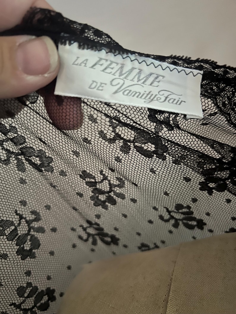 May include: Black lace fabric with a floral pattern. The tag reads "LA FEMME DE Vanity Fair". The lace is delicate and detailed, with a fine mesh background and intricate floral designs. The fabric is likely used for lingerie or other delicate garments.