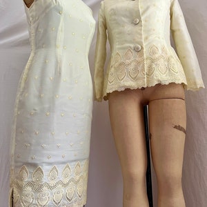 Pode incluir: Um conjunto vintage de duas pe&ccedil;as em cor amarelo claro. O vestido &eacute; sem mangas, ajustado, com gola mandarim e uma delicada renda. A jaqueta tem mangas compridas, gola e uma renda semelhante. Ambas as pe&ccedil;as s&atilde;o adornadas com pequenos pontos bordados.