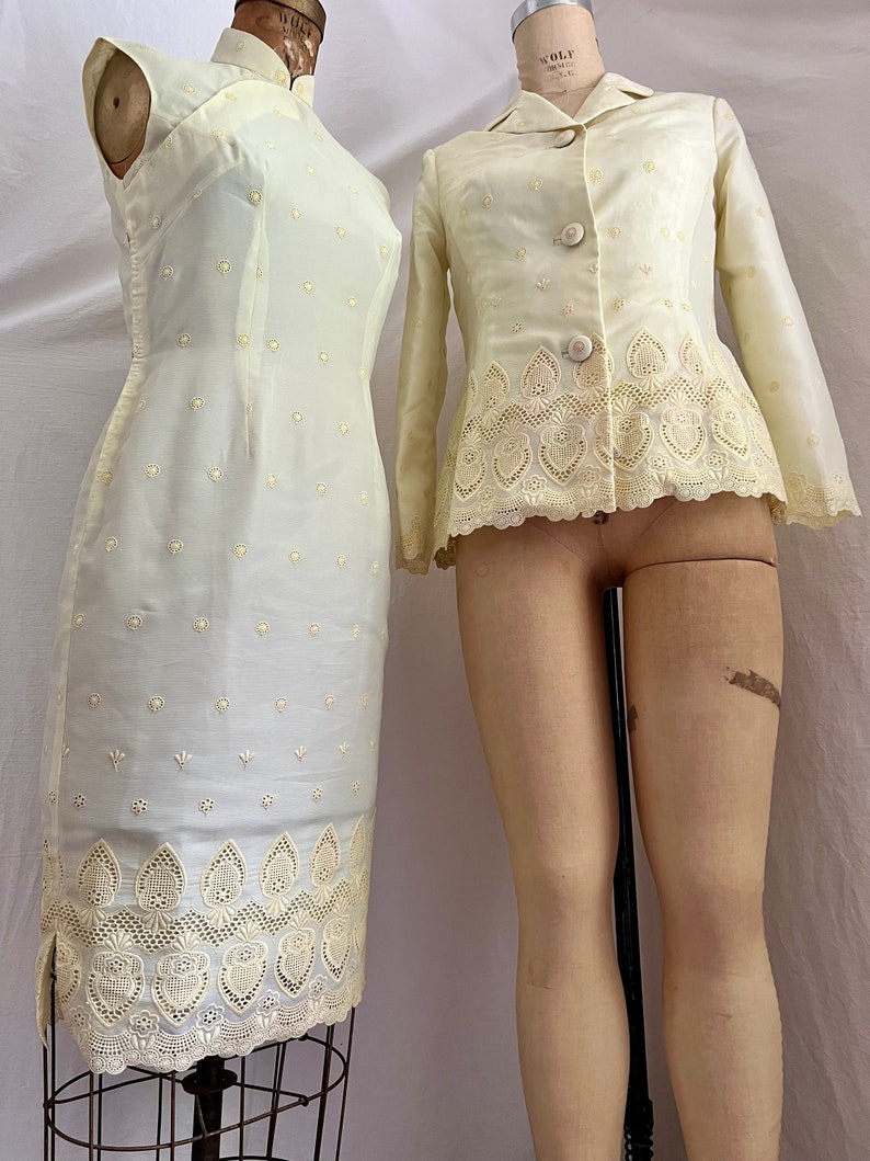 Pode incluir: Um conjunto vintage de duas pe&ccedil;as em cor amarelo claro. O vestido &eacute; sem mangas, ajustado, com gola mandarim e uma delicada renda. A jaqueta tem mangas compridas, gola e uma renda semelhante. Ambas as pe&ccedil;as s&atilde;o adornadas com pequenos pontos bordados.