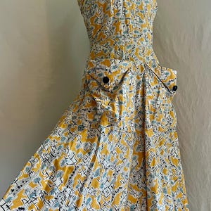 K&ouml;nnte beinhalten: Ein Kleid im Vintage-Stil mit einem gelben, wei&szlig;en und blauen Muster. Das Kleid hat ein figurbetontes Oberteil, einen ausgestellten Rock und zwei Vordertaschen. Es hat schwarze Kn&ouml;pfe und ein tr&auml;gerloses Design.