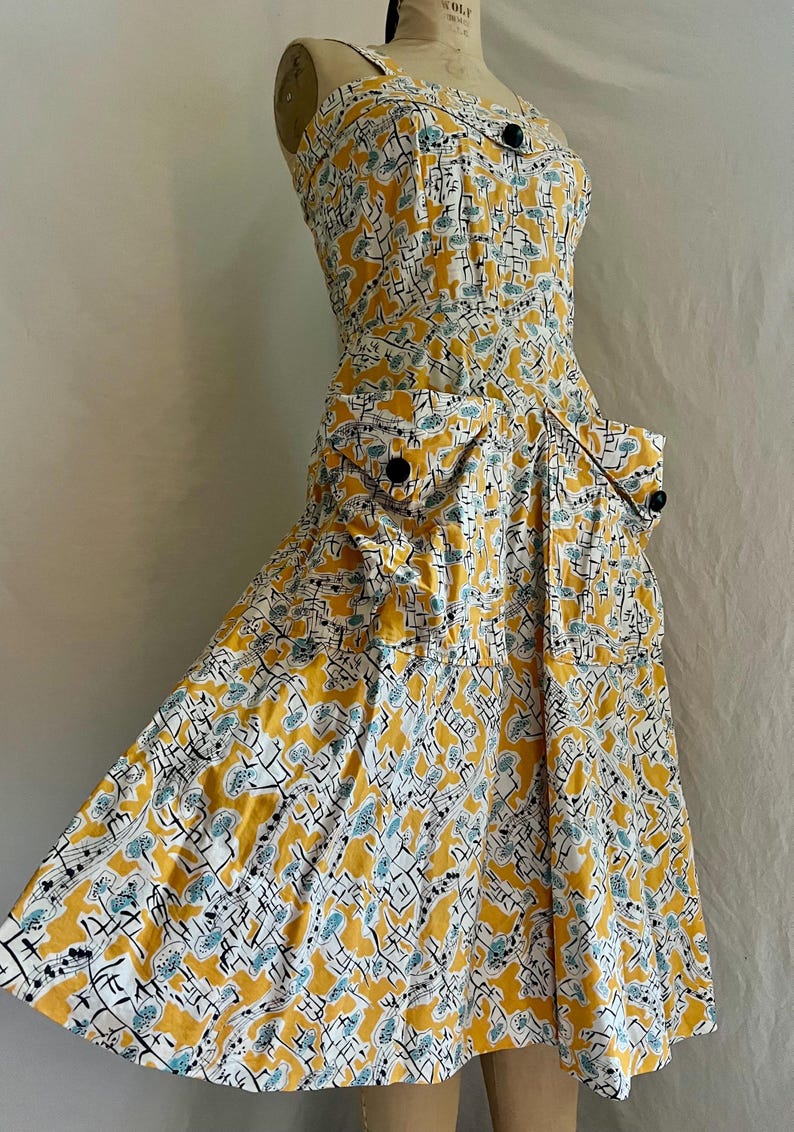 K&ouml;nnte beinhalten: Ein Kleid im Vintage-Stil mit einem gelben, wei&szlig;en und blauen Muster. Das Kleid hat ein figurbetontes Oberteil, einen ausgestellten Rock und zwei Vordertaschen. Es hat schwarze Kn&ouml;pfe und ein tr&auml;gerloses Design.