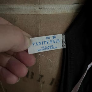 May include: A close-up of a tag from a vintage garment. The tag reads "VANITY FAIR" with the size "30" printed above. The tag also indicates the material is nylon. The garment is black.