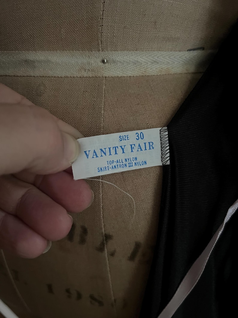 May include: A close-up of a tag from a vintage garment. The tag reads "VANITY FAIR" with the size "30" printed above. The tag also indicates the material is nylon. The garment is black.