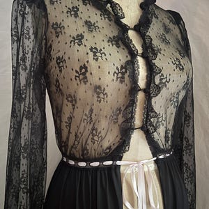 May include: Black lace lingerie set featuring a sheer, long-sleeved top with floral patterns and a ruffled trim. The set includes a matching black skirt and a white ribbon tie at the waist. The garment is displayed on a mannequin.