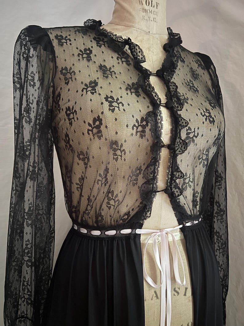 May include: Black lace lingerie set featuring a sheer, long-sleeved top with floral patterns and a ruffled trim. The set includes a matching black skirt and a white ribbon tie at the waist. The garment is displayed on a mannequin.