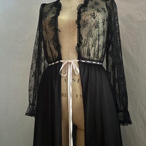 May include: Black vintage lingerie set featuring a sheer lace top with floral patterns and ruffled edges. The long-sleeved top is paired with a solid black, floor-length skirt. A white ribbon cinches the waist, adding a delicate touch to the elegant ensemble.