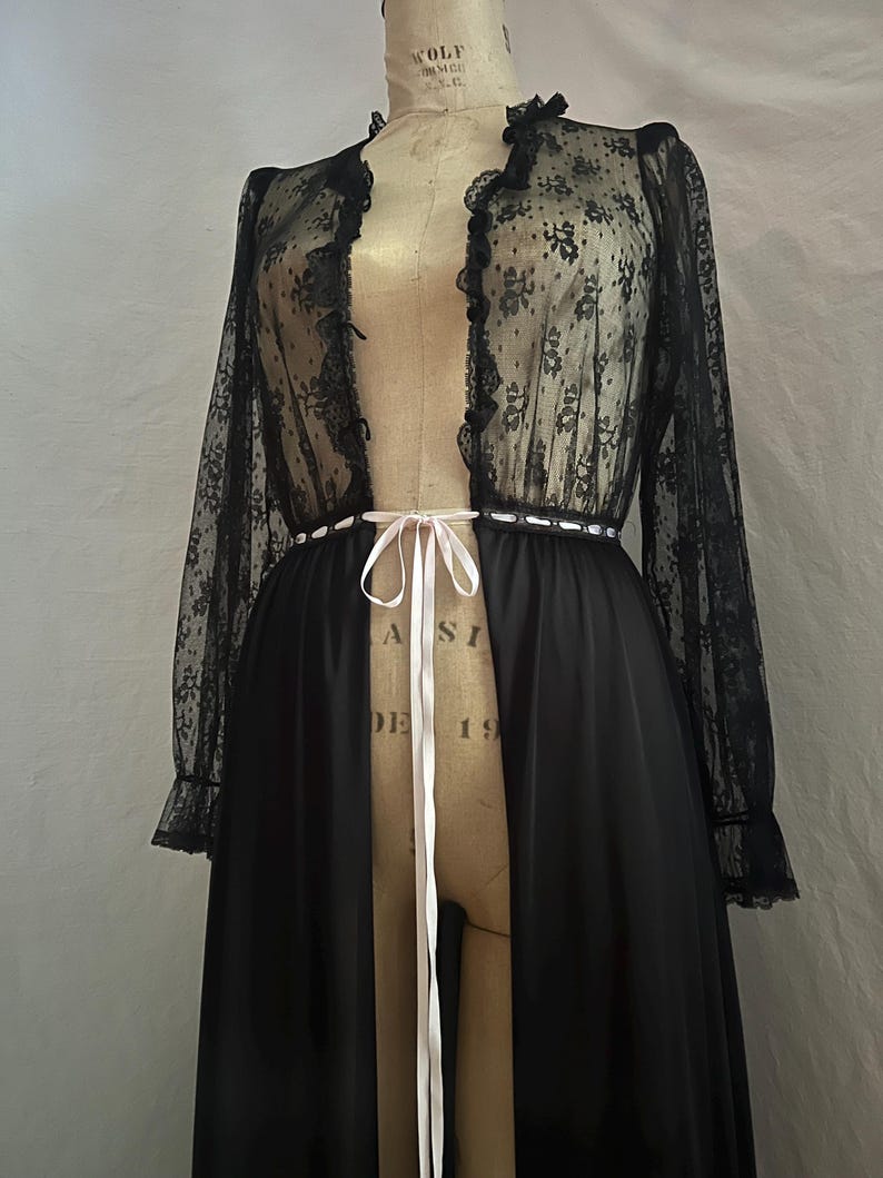 May include: Black vintage lingerie set featuring a sheer lace top with floral patterns and ruffled edges. The long-sleeved top is paired with a solid black, floor-length skirt. A white ribbon cinches the waist, adding a delicate touch to the elegant ensemble.