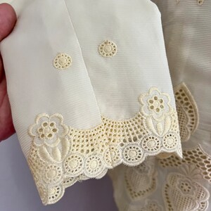 Pode incluir: Uma blusa branca bordada com um delicado padr&atilde;o floral. A blusa tem uma bainha de renda festoneada e um fecho de bot&otilde;es na frente.