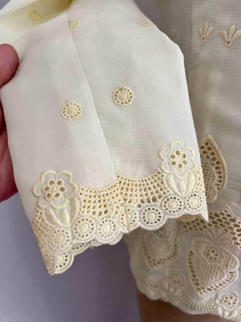 Pode incluir: Uma blusa branca bordada com um delicado padr&atilde;o floral. A blusa tem uma bainha de renda festoneada e um fecho de bot&otilde;es na frente.