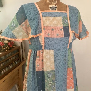 Puede incluir: Un vestido vintage de maxi patchwork con base azul y patrones florales de color melocotón, verde y blanco. El vestido tiene un escote cuadrado y mangas cortas con forma de capucha con un ribete de melocotón.