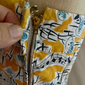 K&ouml;nnte beinhalten: Nahaufnahme eines Vintage-Kleidoberteils mit Messingrei&szlig;verschluss. Der Stoff ist wei&szlig; mit einem sich wiederholenden Muster aus gelben Formen, Noten und blauen Akzenten. Das Kleid befindet sich auf einer Schaufensterpuppe.