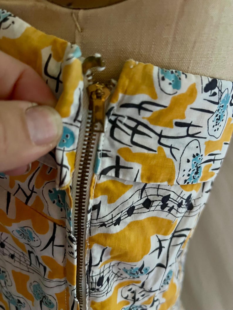 K&ouml;nnte beinhalten: Nahaufnahme eines Vintage-Kleidoberteils mit Messingrei&szlig;verschluss. Der Stoff ist wei&szlig; mit einem sich wiederholenden Muster aus gelben Formen, Noten und blauen Akzenten. Das Kleid befindet sich auf einer Schaufensterpuppe.