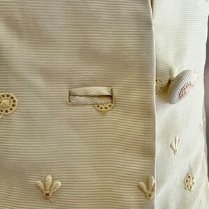 Pode incluir: Um close-up de uma jaqueta branca com tecido texturizado e delicados desenhos florais bordados. A jaqueta tem fecho de bot&atilde;o e um &uacute;nico bolso.