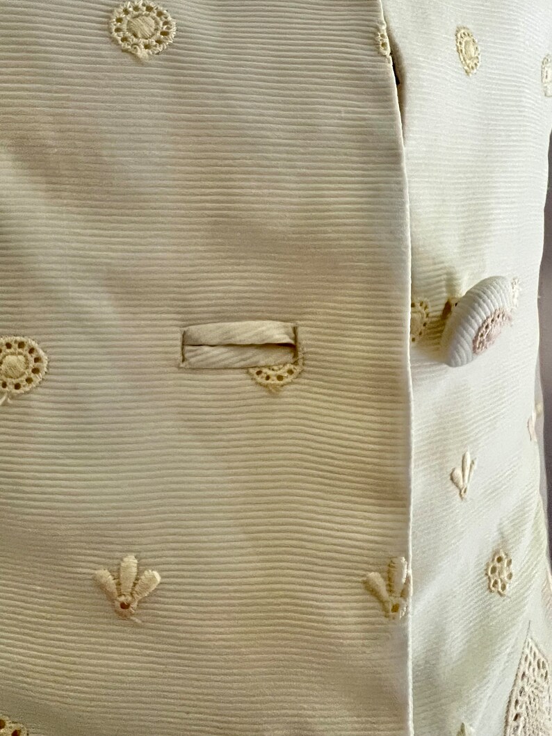 Pode incluir: Um close-up de uma jaqueta branca com tecido texturizado e delicados desenhos florais bordados. A jaqueta tem fecho de bot&atilde;o e um &uacute;nico bolso.