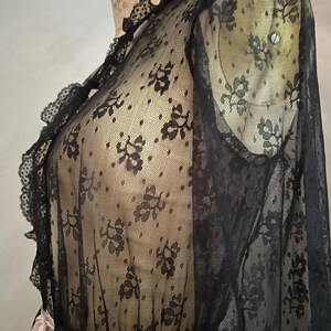 May include: Black lace top with floral patterns and ruffled trim. The sheer fabric drapes over a black garment, with a contrasting white and pink ribbon detail at the waist. The top is displayed on a mannequin.