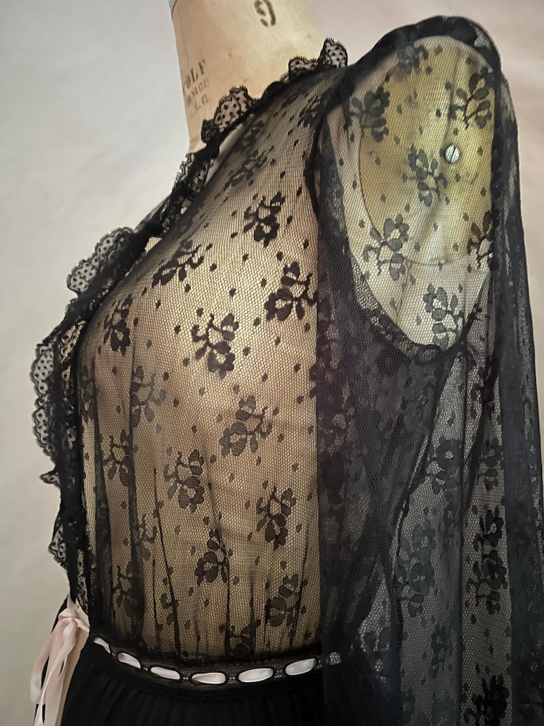 May include: Black lace top with floral patterns and ruffled trim. The sheer fabric drapes over a black garment, with a contrasting white and pink ribbon detail at the waist. The top is displayed on a mannequin.