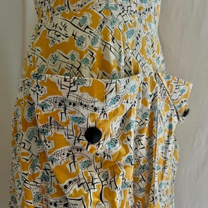 K&ouml;nnte beinhalten: Ein Kleid im Vintage-Stil mit einem einzigartigen Muster. Das Kleid hat einen wei&szlig;en Hintergrund mit einem sich wiederholenden Design aus gelben Formen, schwarzen Linien und t&uuml;rkisfarbenen Akzenten. Es hat gro&szlig;e Taschen mit schwarzen Kn&ouml;pfen und ein eng anliegendes Oberteil.
