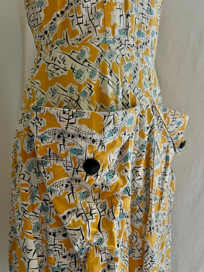K&ouml;nnte beinhalten: Ein Kleid im Vintage-Stil mit einem einzigartigen Muster. Das Kleid hat einen wei&szlig;en Hintergrund mit einem sich wiederholenden Design aus gelben Formen, schwarzen Linien und t&uuml;rkisfarbenen Akzenten. Es hat gro&szlig;e Taschen mit schwarzen Kn&ouml;pfen und ein eng anliegendes Oberteil.