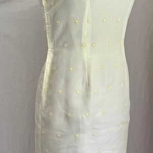 Pode incluir: Um vestido branco sem mangas com gola mandarim e um delicado padr&atilde;o floral. O vestido tem uma silhueta ajustada e uma bainha de renda festonada.