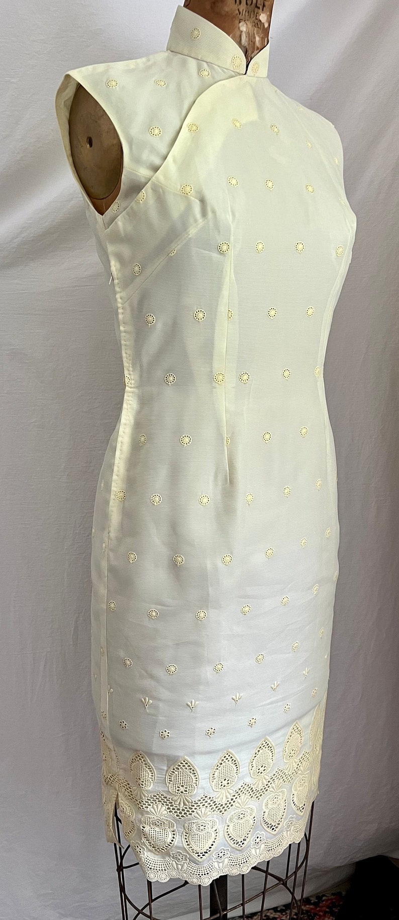 Pode incluir: Um vestido branco sem mangas com gola mandarim e um delicado padr&atilde;o floral. O vestido tem uma silhueta ajustada e uma bainha de renda festonada.