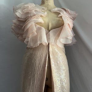 May include: A pale pink evening gown on a dress form. The gown features a sequined skirt with a high slit and a layered, ruffled bodice. The dress is designed for formal occasions.