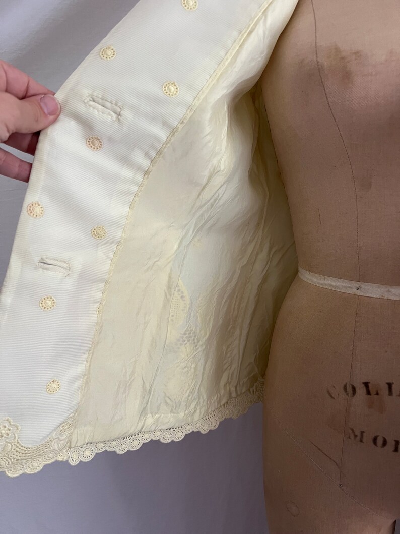 Pode incluir: Uma jaqueta branca com acabamento em renda e detalhes de bot&otilde;es. A jaqueta &eacute; forrada com um tecido branco.