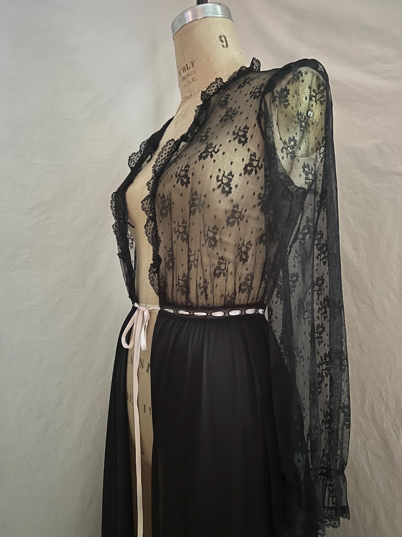 May include: Black lace robe with floral patterns and long sleeves. The robe has a sheer design with a black skirt and a white ribbon belt. The robe is displayed on a mannequin.