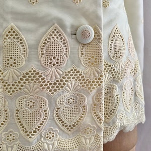 Pode incluir: Uma jaqueta branca bordada com uma bainha festonada e um fecho de bot&atilde;o &uacute;nico. O bordado apresenta um delicado padr&atilde;o floral e um design de cora&ccedil;&atilde;o.