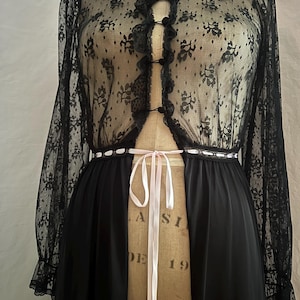 May include: Black vintage lingerie set featuring a sheer lace top with floral patterns, long sleeves, and a ruffled collar. The set includes a long, flowing black skirt, and a pink ribbon tie at the waist. The garment is displayed on a vintage dress form.