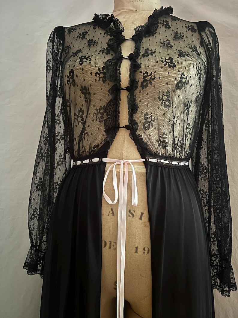 May include: Black vintage lingerie set featuring a sheer lace top with floral patterns, long sleeves, and a ruffled collar. The set includes a long, flowing black skirt, and a pink ribbon tie at the waist. The garment is displayed on a vintage dress form.