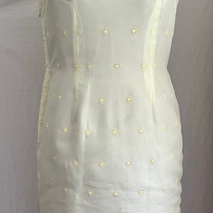 Pode incluir: Um vestido branco sem mangas com gola mandarim e uma bainha de renda festonada. O vestido &eacute; coberto por pequenos c&iacute;rculos bordados brancos.